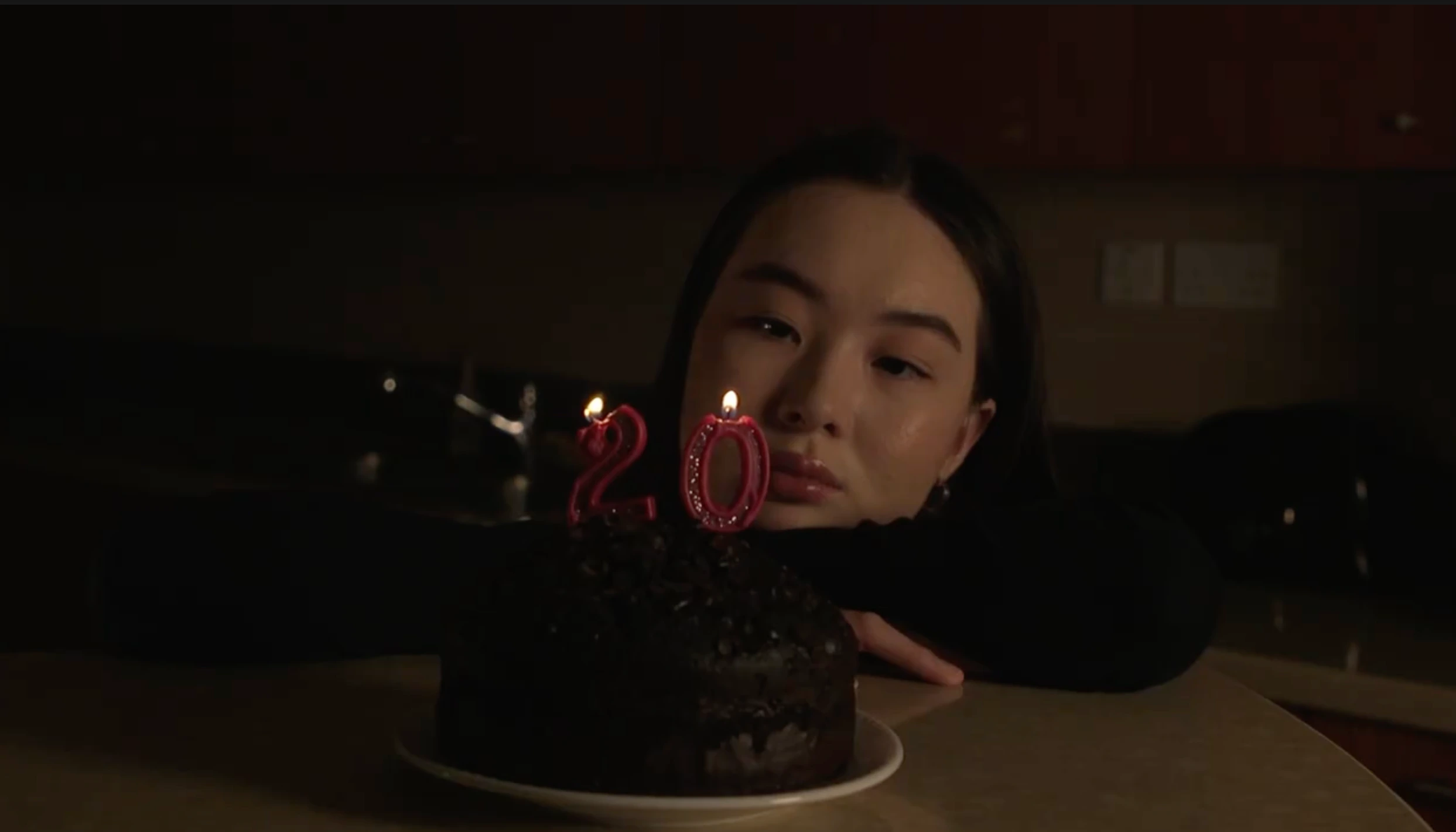 A young woman rests beside a chocolate birthday cake with burning number-20 candles in a dim room.