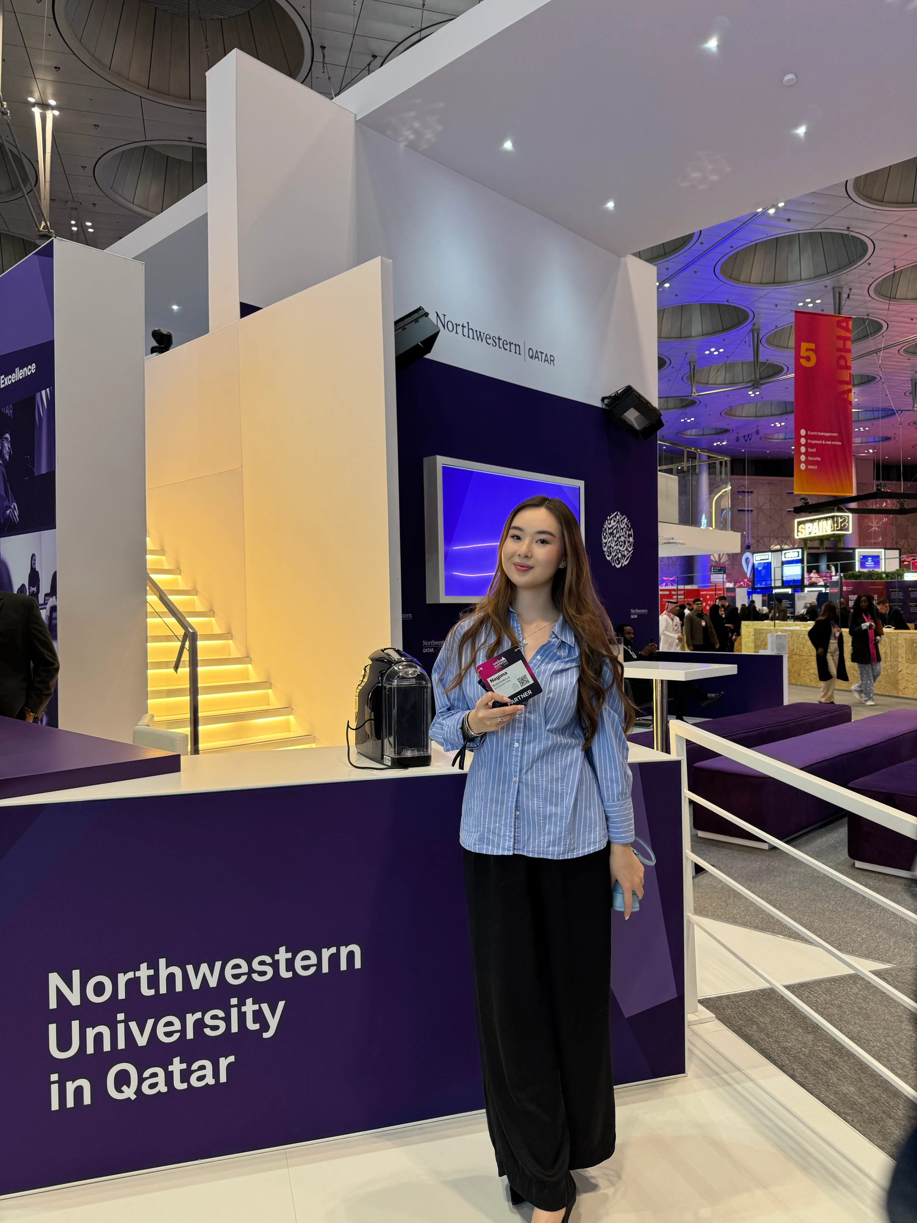 Nagima Bolotbekova stands beside a Northwestern University in Qatar exhibition stand, holding an event badge.